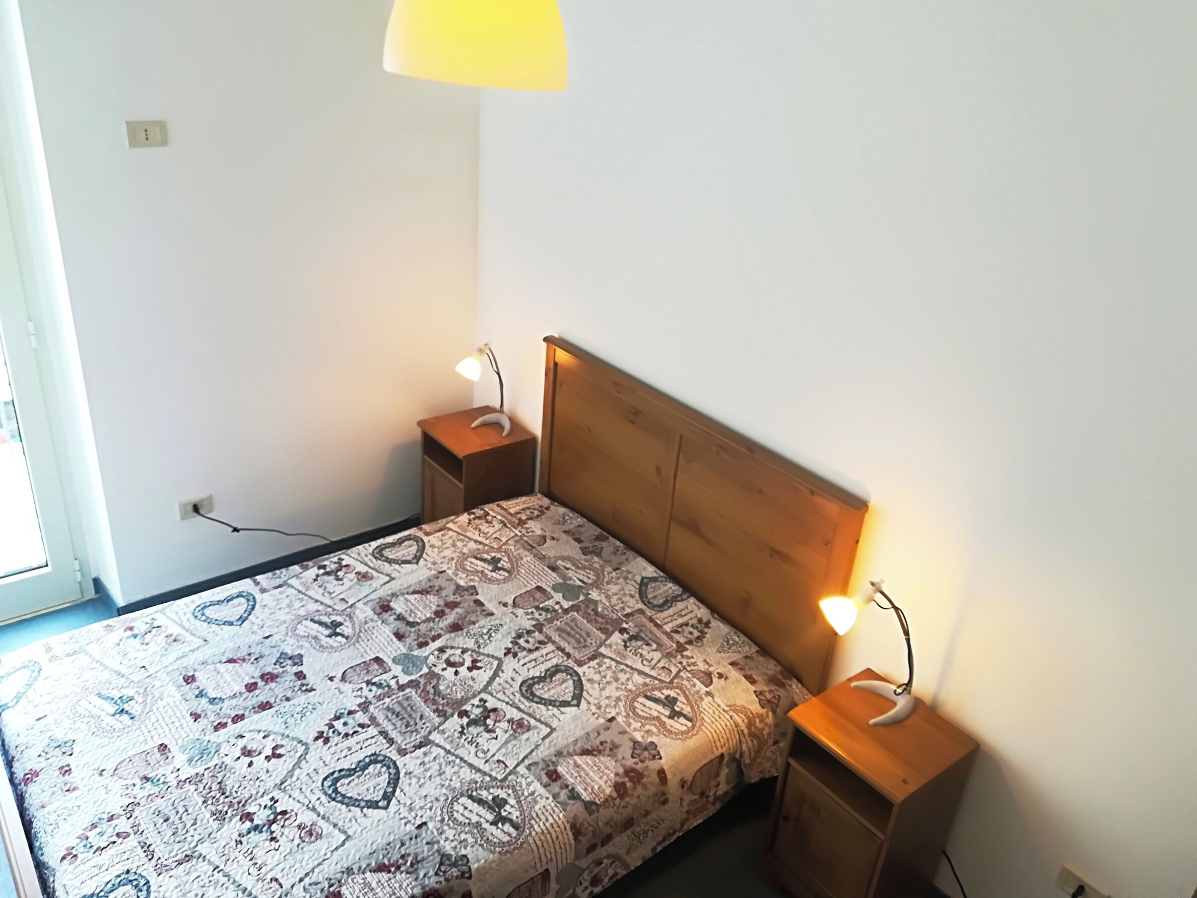 Makuuhuoneessa on puinen sängynpääty, kuviollinen päiväpeite, kaksi yöpöytää, joissa on lamput, ja ikkuna, josta on näkymä.