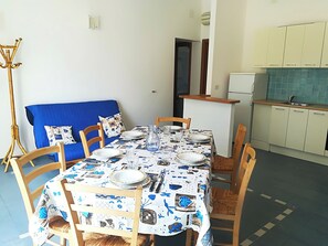 Ruokailuhuone, jossa on neljälle katettu ruokapöytä, tuolit, sininen sohva sekä keittiöalue, jossa on kaappeja ja jääkaappi.