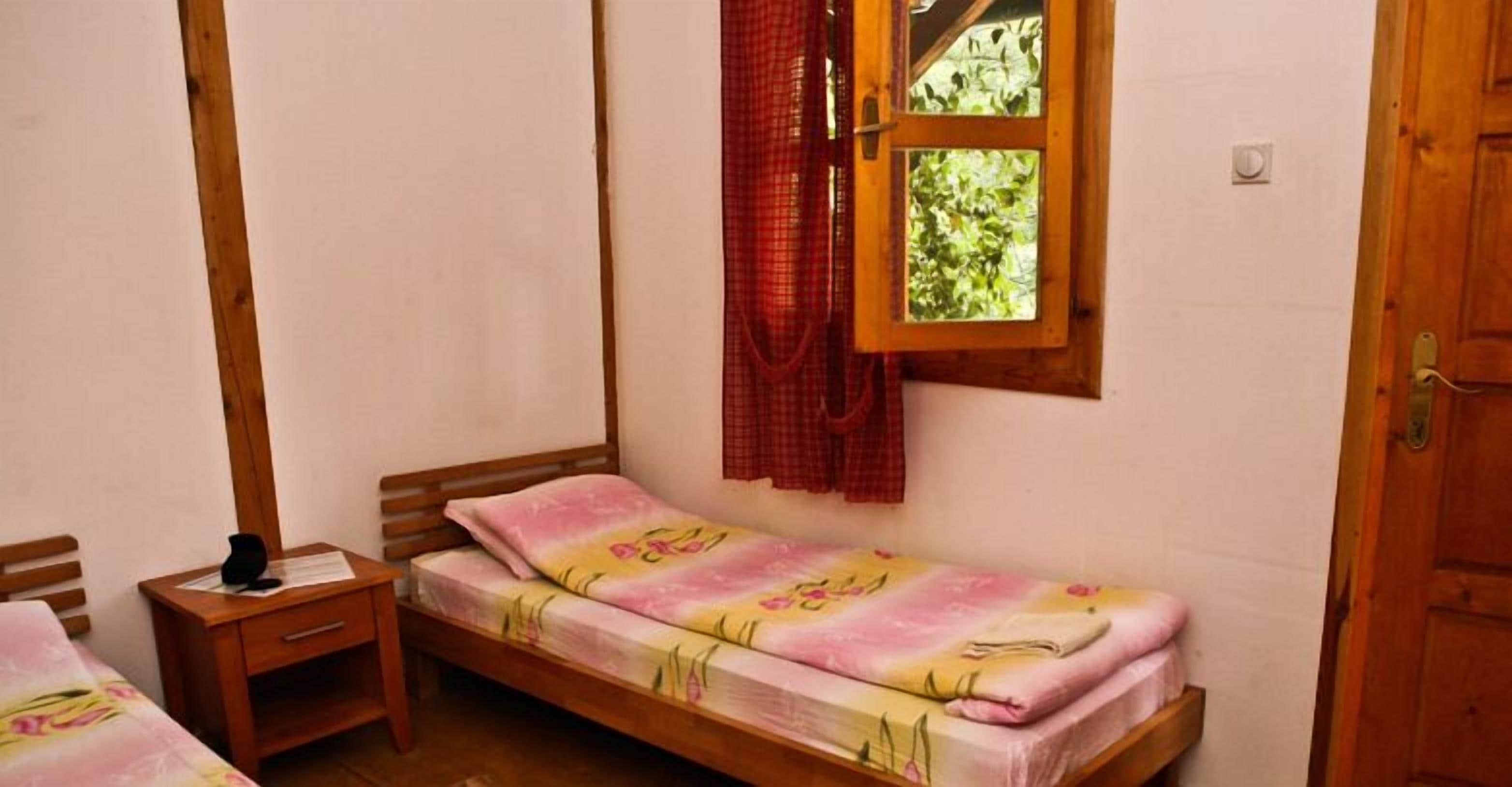 A single bed with a floral bedspread, a wooden nightstand, a window with a red curtain, and a wooden door.