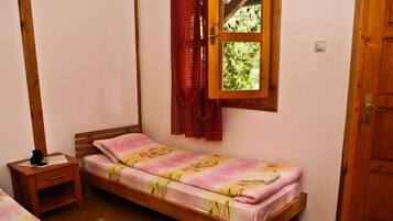 A single bed with a floral bedspread, a wooden nightstand, a window with a red curtain, and a wooden door.