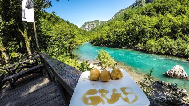 A wooden deck with a white plate of food and a flag, overlooking a turquoise river and green hills.