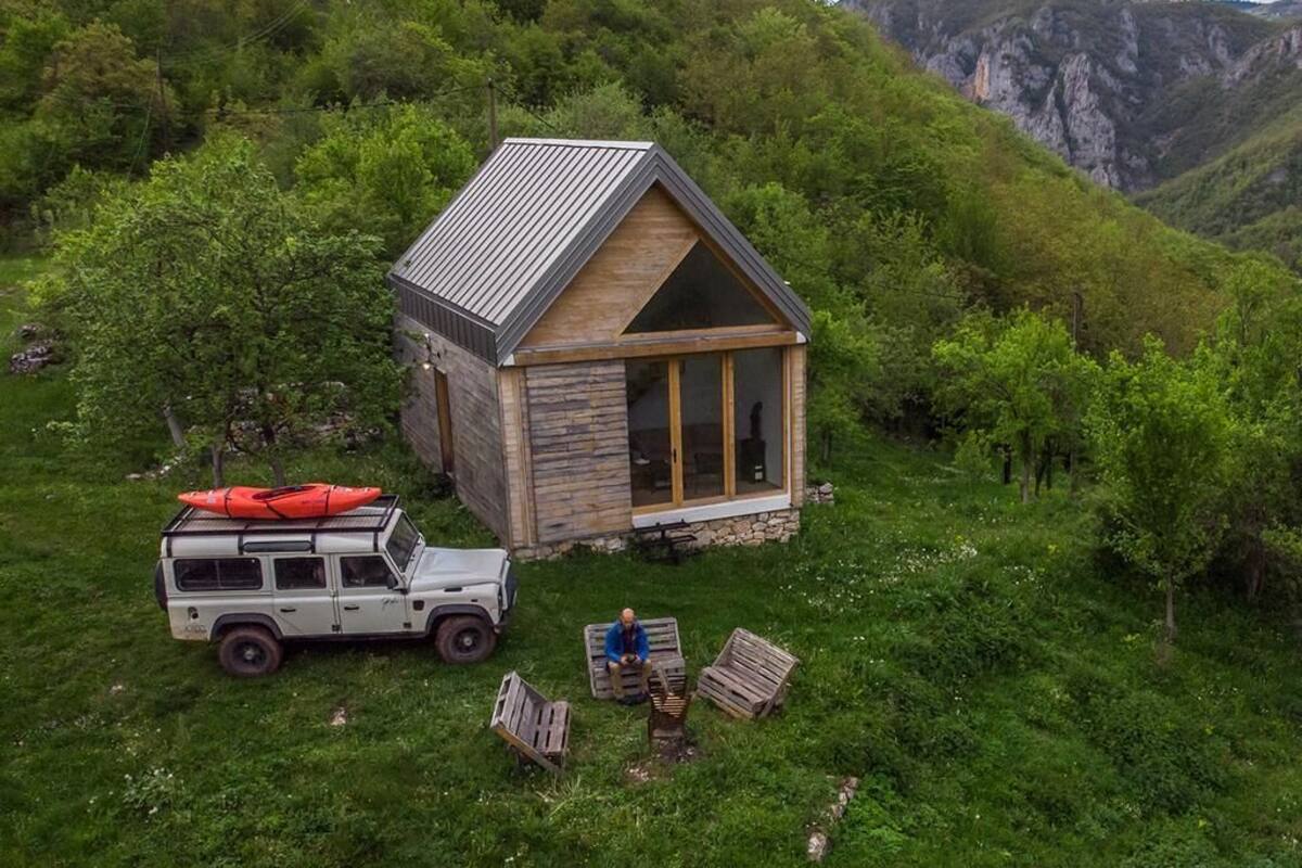 A small cabin with a sloped roof, surrounded by lush greenery and mountains.