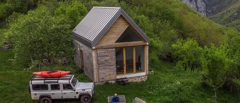 A small cabin with a sloped roof, surrounded by lush greenery and mountains.