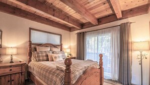 Quarto com cama de madeira, mesa de cabeceira, abajur, janela com persianas e vista para o exterior.