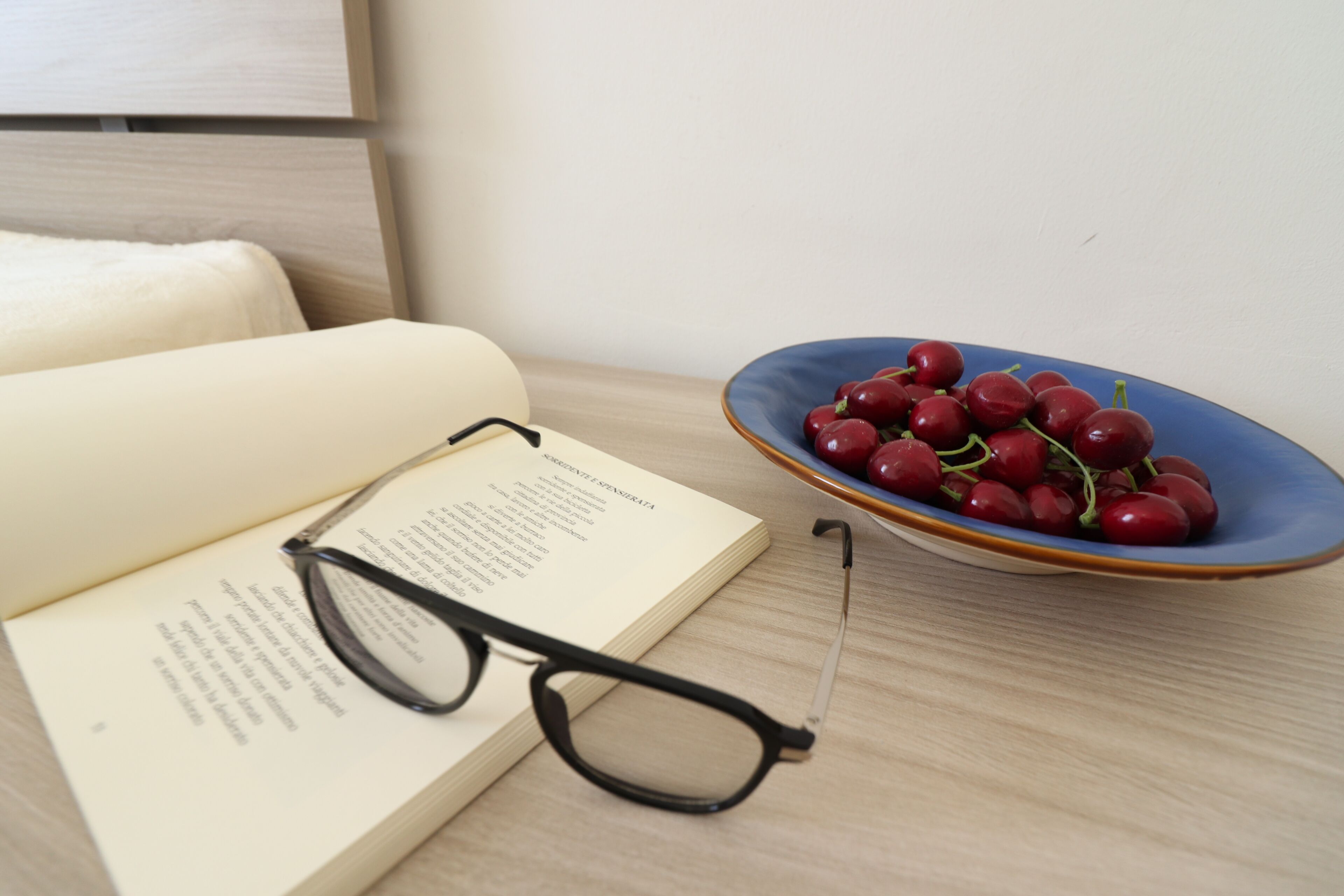 A book, a pair of glasses, and a plate of cherries on a wooden table.