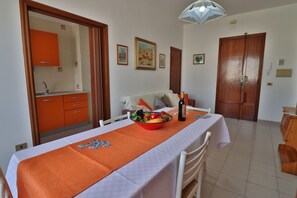 A dining area with a table set for two, a fruit bowl, and a bottle of wine. There is a sofa and a wooden door in the background. - Casa Vacanza Atena Otranto 6 Posti (Otranto)