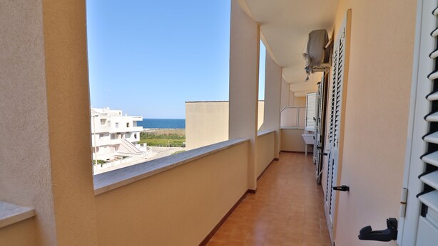 Un pasillo con ventana que da a una playa y edificios, que conduce a una puerta con ventana y un pequeño balcón.