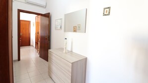 Un pasillo con una puerta de madera, suelo de baldosas blancas, una consola de color claro con un florero y cuadros en la pared.