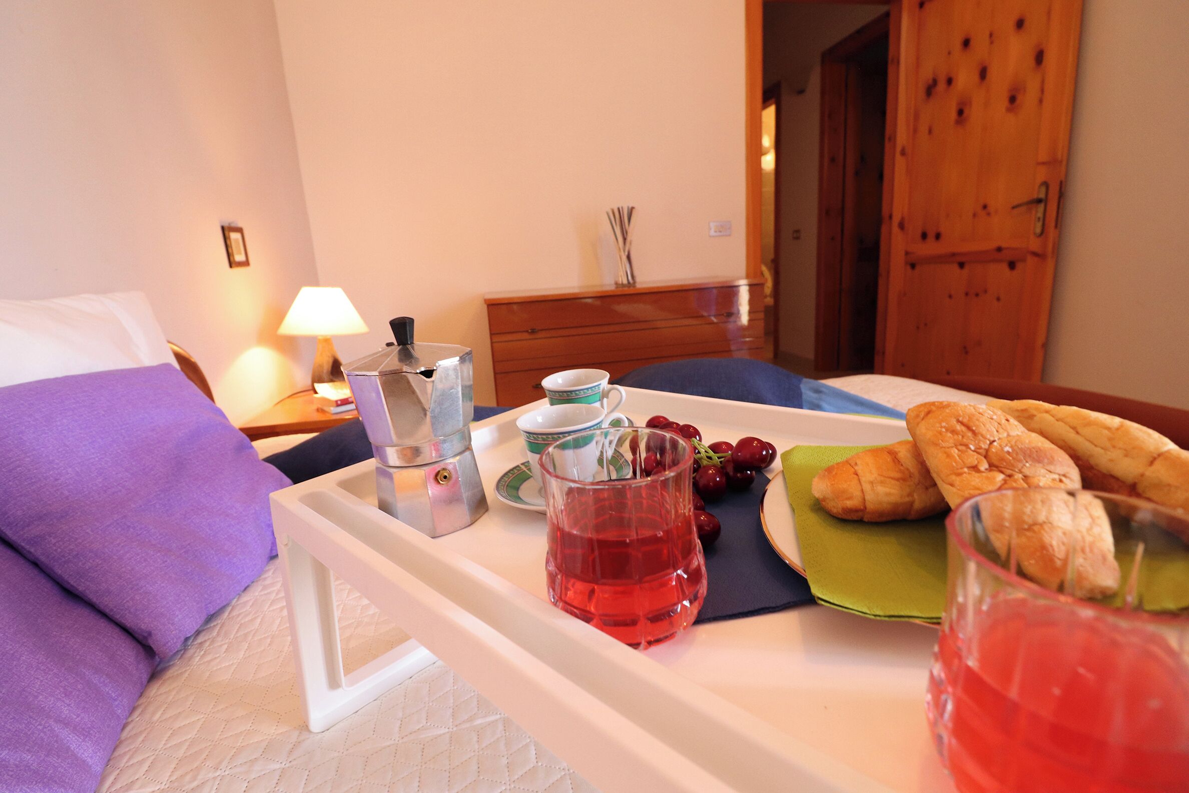 Ein ordentlich bezogenes Bett mit einem violetten Kopfkissen, ein Nachttisch mit einer Lampe, ein Tablett mit Speisen und Getränken und eine Holztür im Hintergrund.