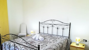 Un lit simple avec un cadre en métal, une couvre-lit à motifs, deux serviettes enroulées, une table de chevet avec une lampe et une chaise.