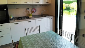 Une cuisine compacte équipée d’une cuisinière à gaz, d’un évier et de meubles blancs. On y trouve une table à manger recouverte d ’une nappe à motifs, ainsi qu’une chaise.
