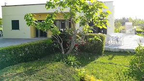 Un bâtiment moderne doté d’un toit plat, d’un petit jardin avec un arbre et des arbustes, et d’un espace pavé aménagé avec du mobilier d’extérieur.