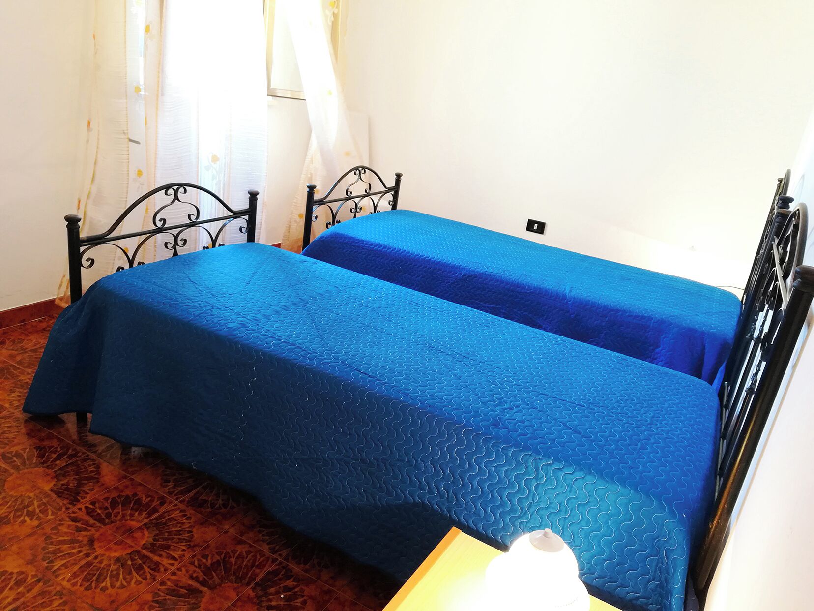 A room with two beds, each covered with blue bedding. The room has a wooden floor and a white wall.