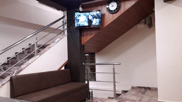 A lobby with a brown sofa, a staircase, a clock, and a television mounted on the wall. - Punest Hospitality & Hotel (Pune)