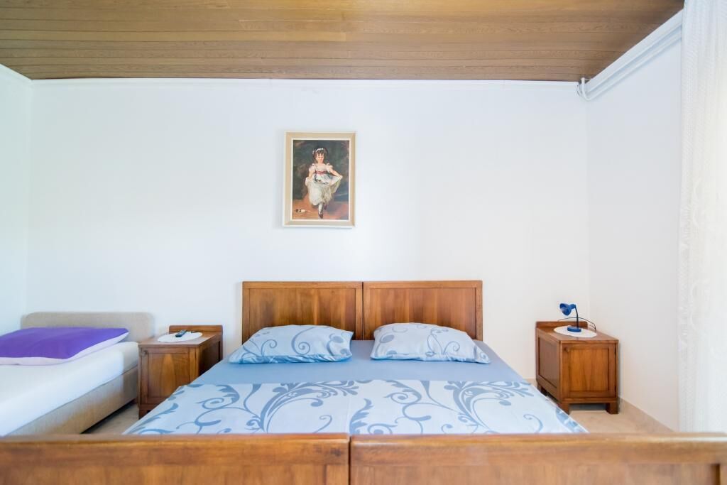 Une chambre à coucher avec un lit en bois, deux tables de chevet, une commode, un tableau au mur et une fenêtre avec des rideaux blancs.