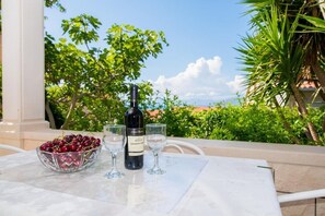 Une table avec une bouteille de vin, des verres et un bol de cerises, sur une terrasse offrant une vue sur la verdure et les bâtiments.