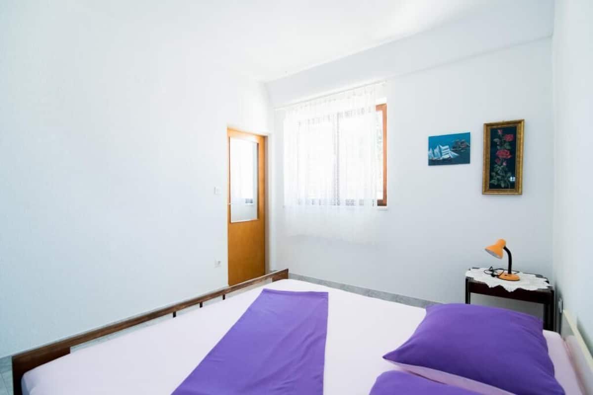 A room with a bed, a door, a window with curtains, a small table with a lamp, and two framed pictures on the wall.