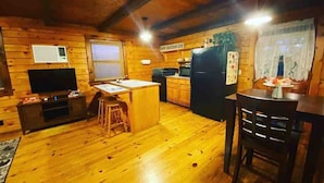 Interior acogedor de una cabaña con paredes de madera, una zona de cocina, una mesa de comedor y un televisor.