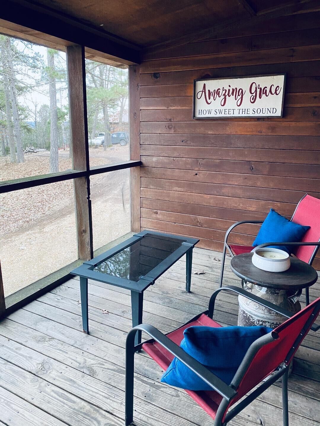 A wooden deck with a glass table and chairs, a sign reading "Amazing Grace," and a view of trees and a road.