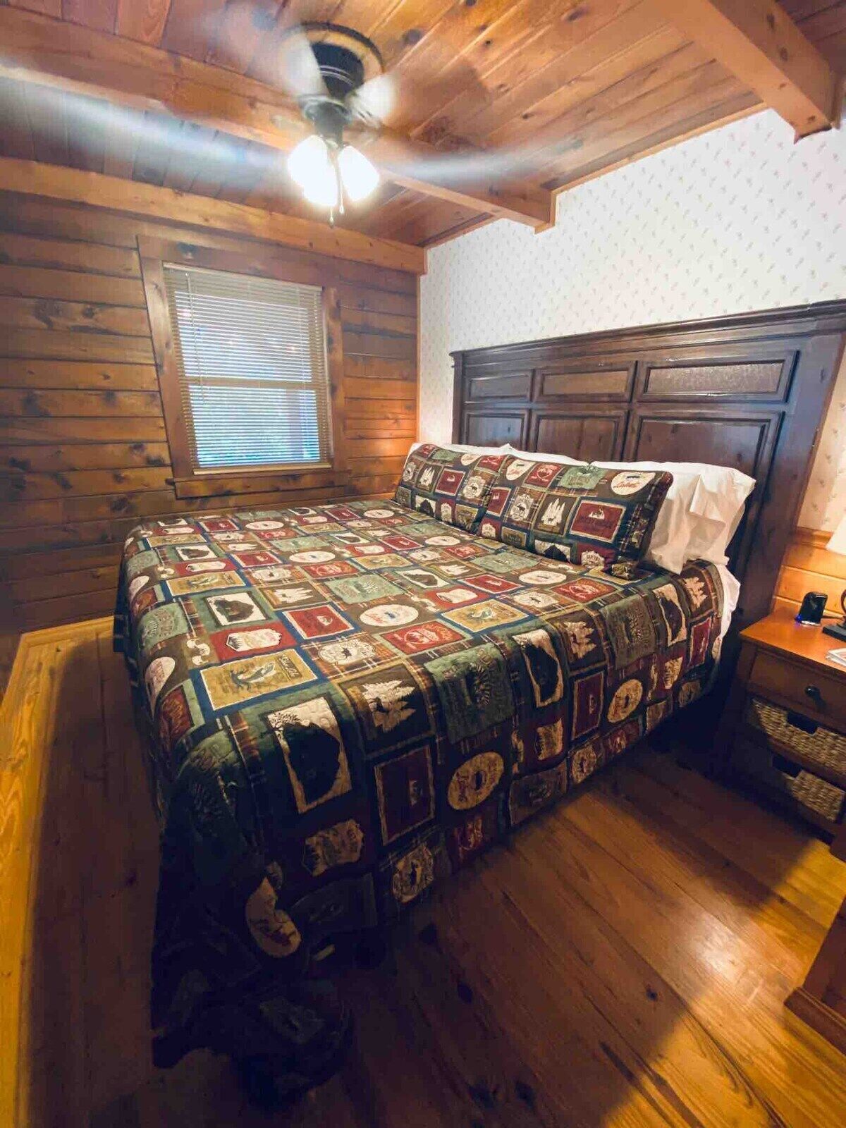 A cozy bedroom with a wooden ceiling, a quilted bedspread, a wooden headboard, and a window with blinds.