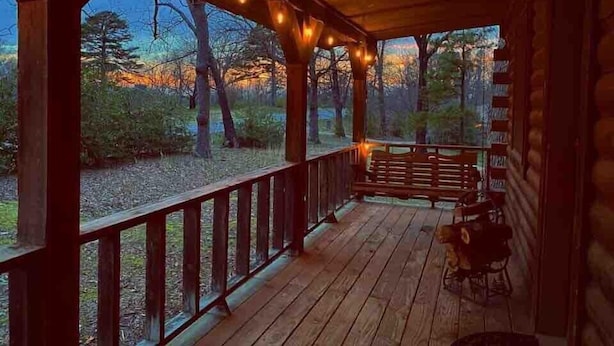 Un porche de madera con luces colgantes, un banco y vistas a árboles y un atardecer.