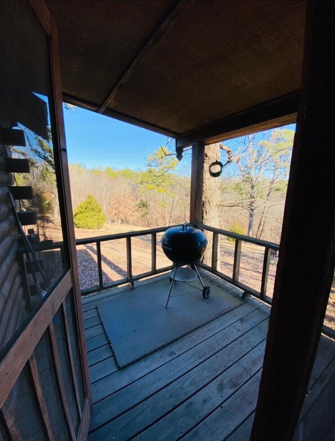 A covered porch with a grill, wooden deck, and a view of trees and sky.