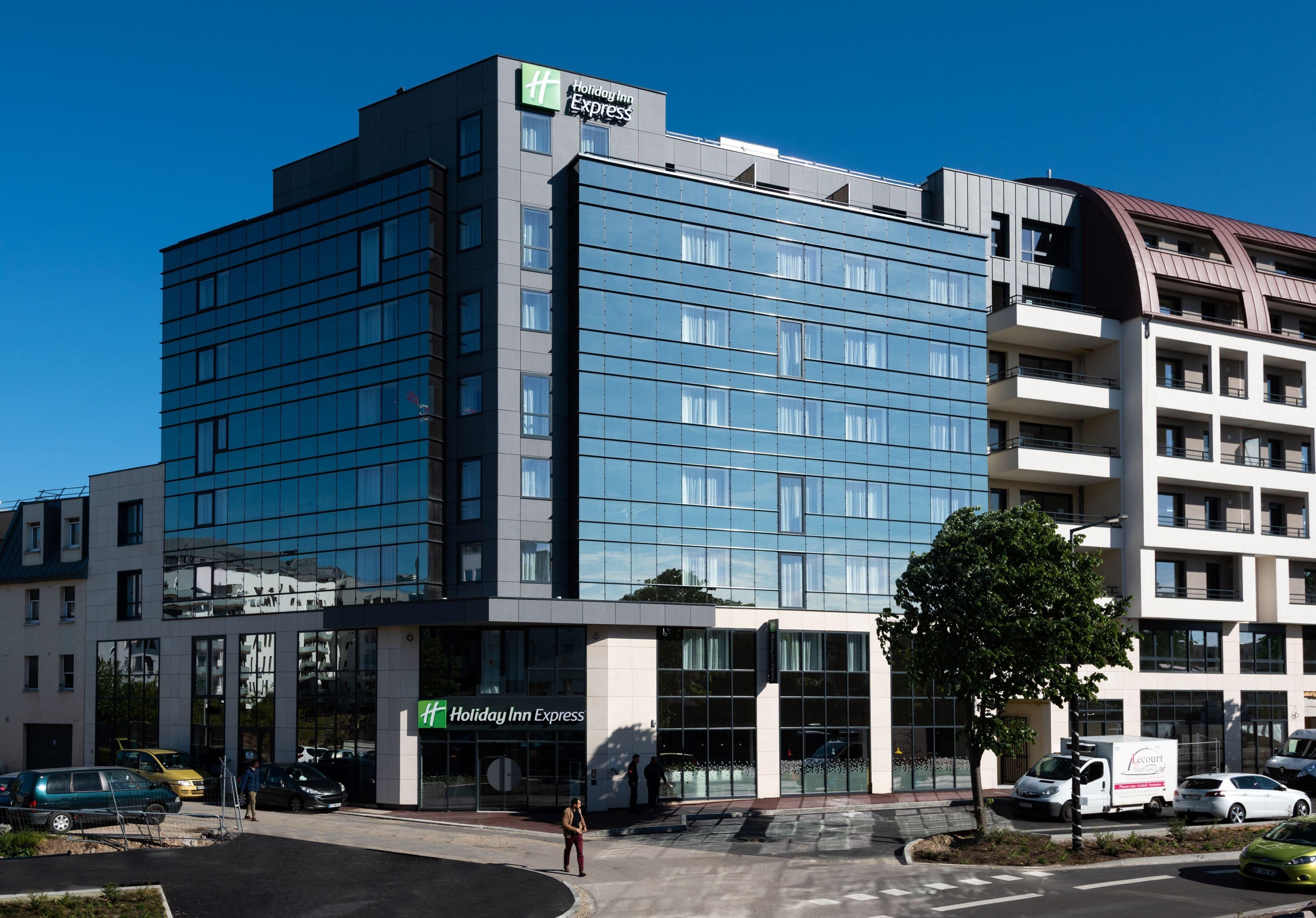 A modern Holiday Inn Express building with a glass facade, multiple floors, and a parking area with cars.