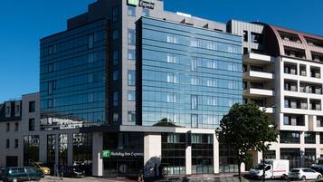 A modern Holiday Inn Express building with a glass facade, multiple floors, and a parking area with cars.