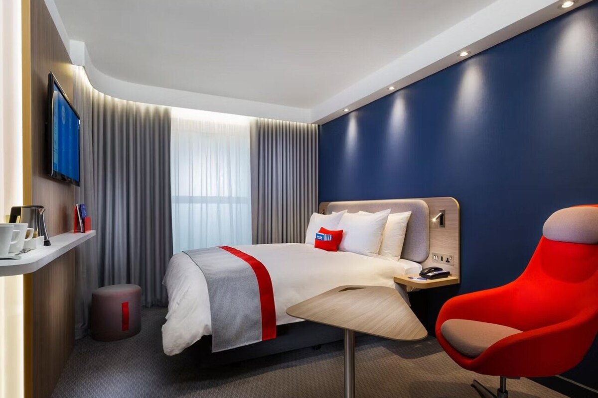Een moderne hotelkamer met een bed, een rode stoel, een bureau en een televisie aan de muur.