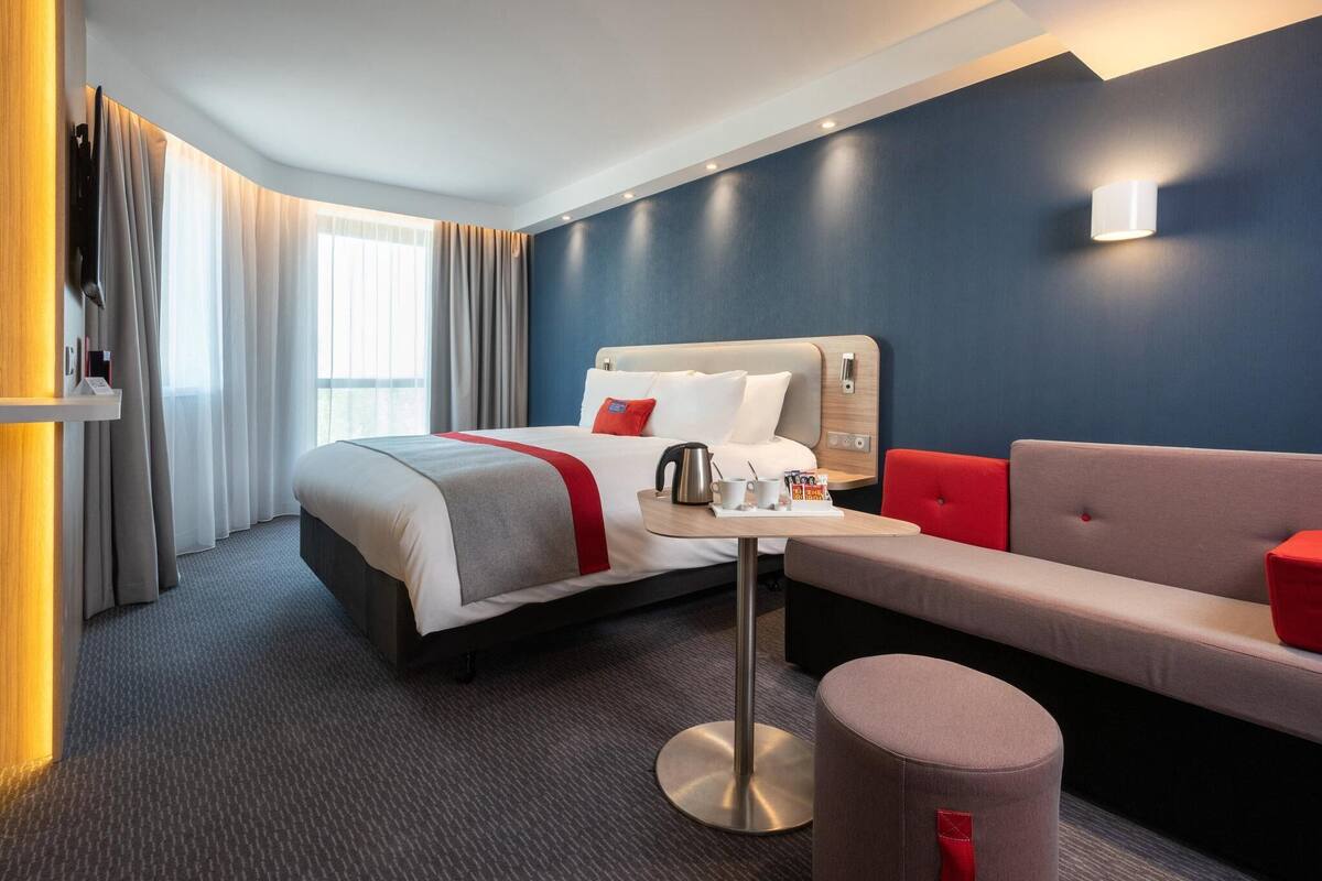 A hotel room with a bed, a sofa, a small table, and a trash bin.
