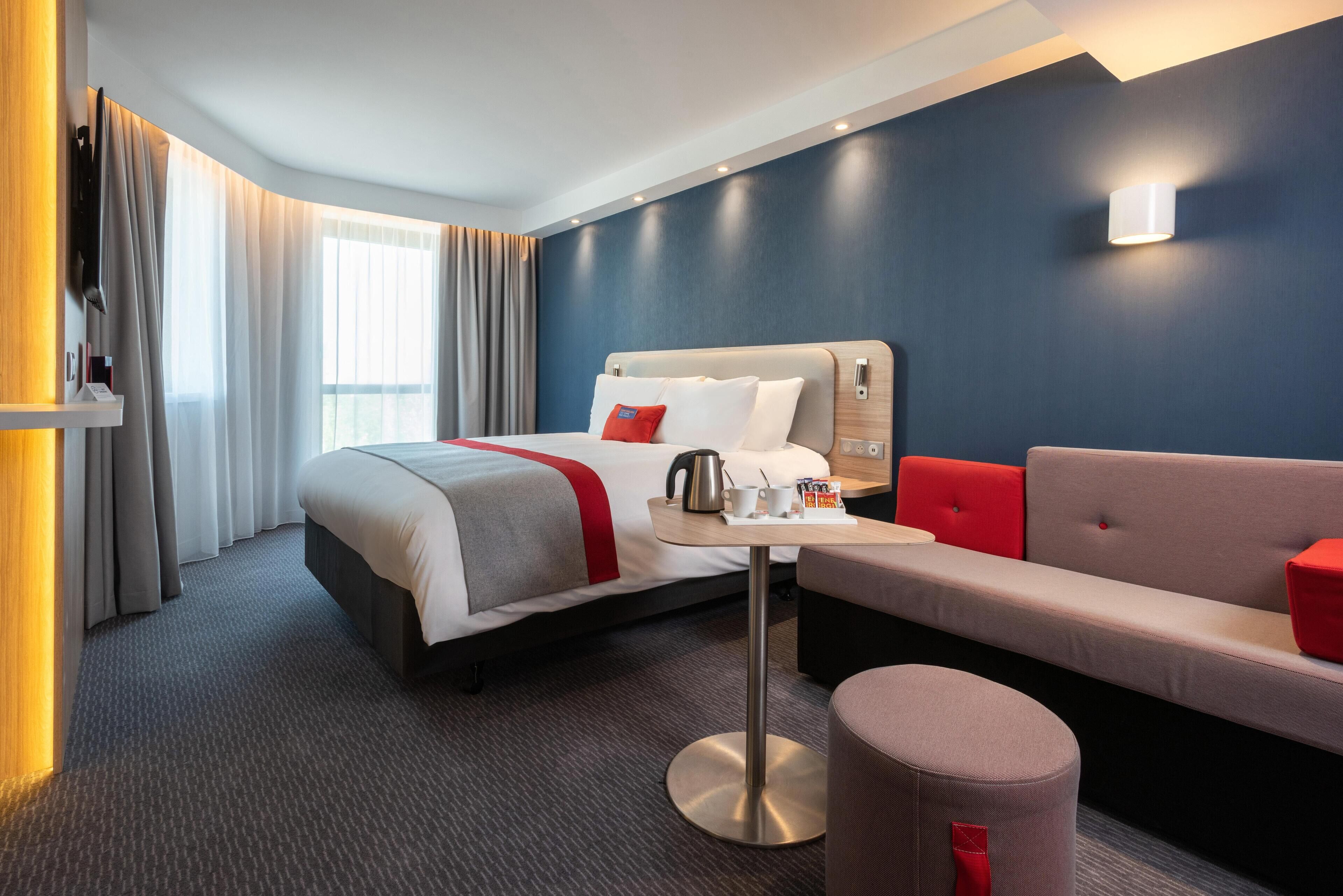 Quarto de hotel com cama, uma pequena mesa com chaleira, um sofá e uma almofada decorativa vermelha.