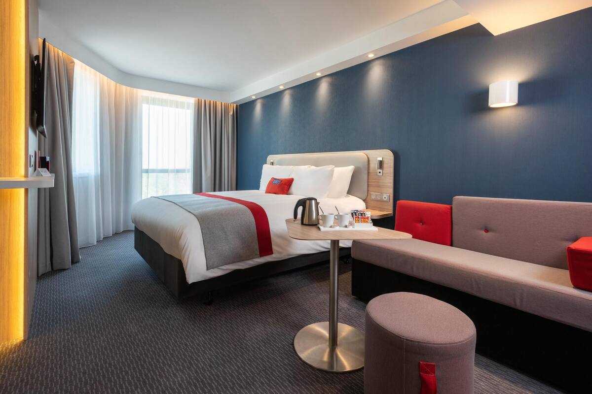 A hotel room with a bed, a small table with a kettle, a sofa, and a red accent pillow.