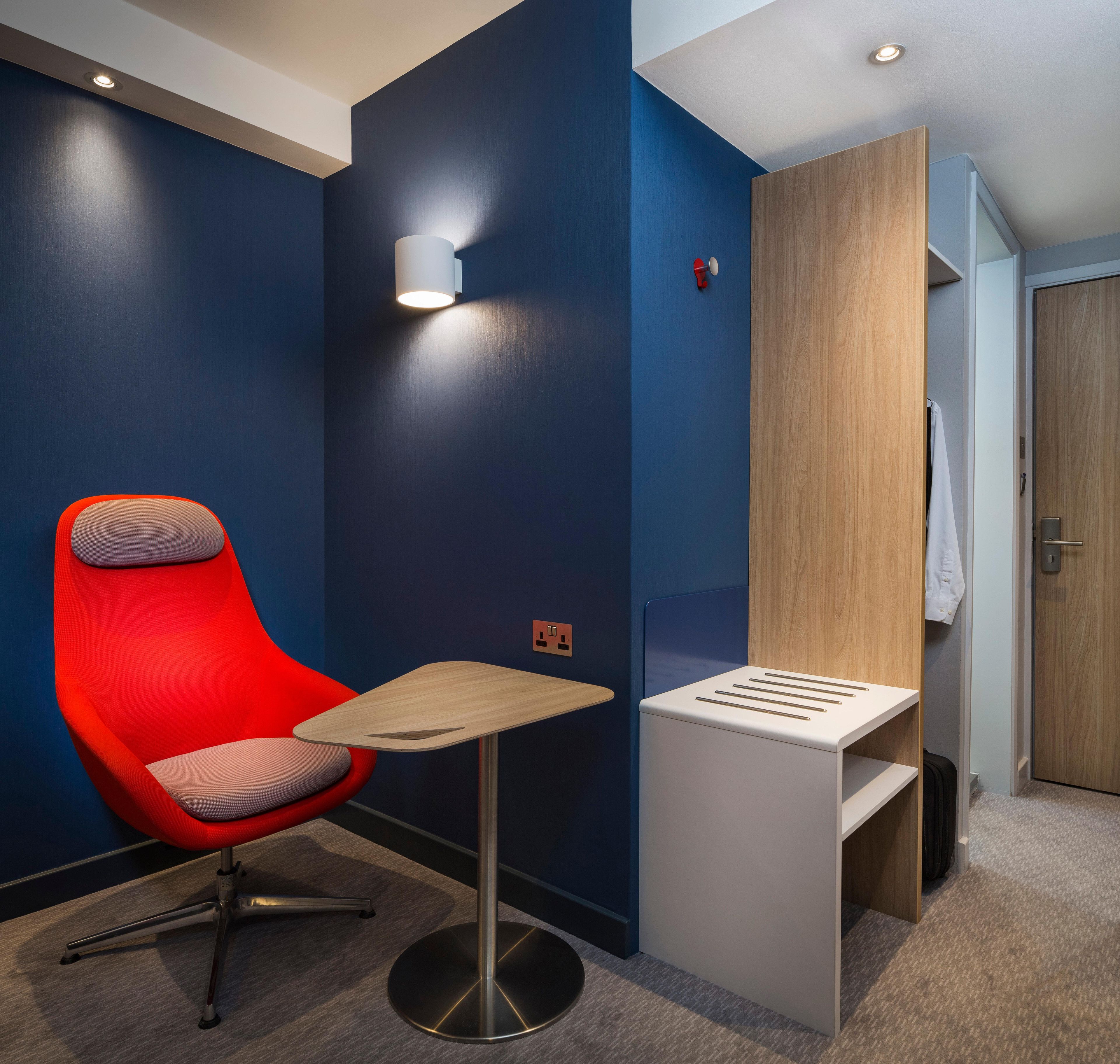 Una habitación de hotel moderna con una silla roja, una mesita pequeña y un armario de madera.