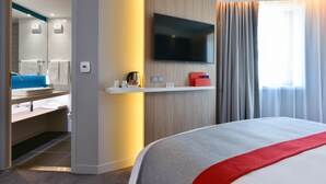 Quarto de hotel moderno com uma cama grande, televisão na parede, uma pequena prateleira com artigos de higiene e um banheiro visível através de uma porta aberta.