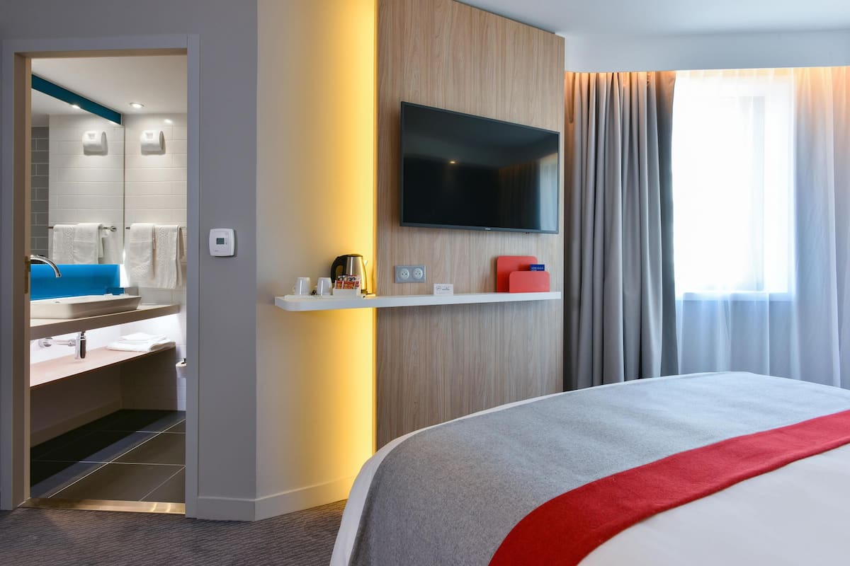 A modern hotel room with a large bed, a mounted TV, a small shelf with toiletries, and a bathroom visible through an open door.
