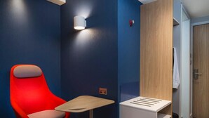 Quarto de hotel moderno com uma cadeira vermelha, uma mesa pequena e um banco embutido com um candeeiro em cima.