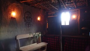 A dimly lit room with a wooden bench, a wall-mounted plant, and a window with a red checkered curtain. - Olonyori Maasai House (Maasai Mara)