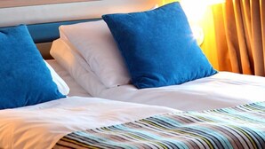 Una habitación de hotel con una cama, ropa de cama blanca y azul, una manta a rayas y un cabecero con detalles de madera.