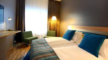 Une chambre d’hôtel avec un lit, deux chaises, un bureau, une lampe et une fenêtre avec des rideaux.