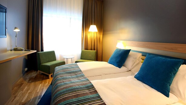 Una habitación de hotel con cama, dos sillas, escritorio, lámpara y ventana con cortinas.