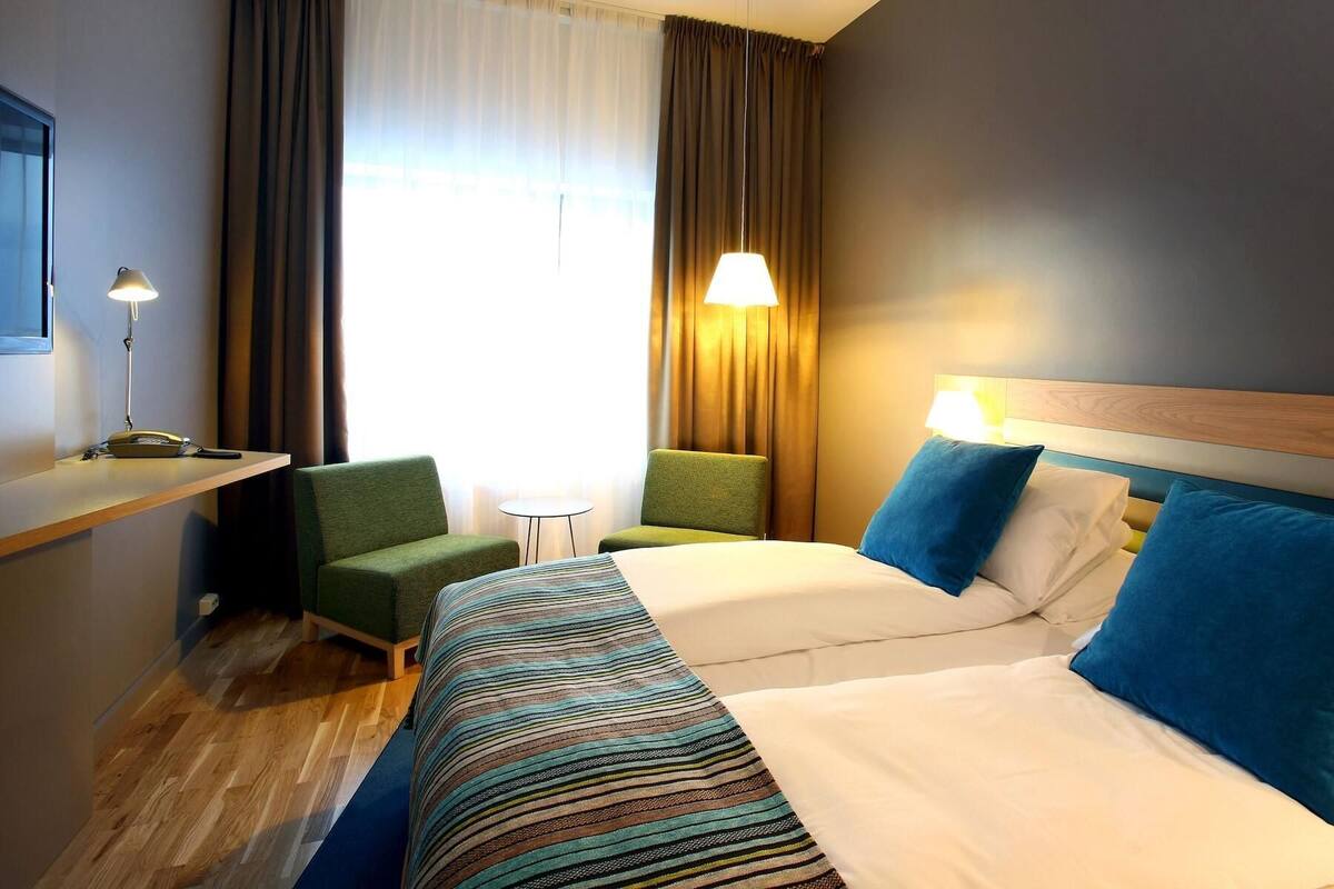 Een hotelkamer met een bed, twee stoelen, een bureau, een lamp en een raam met gordijnen.