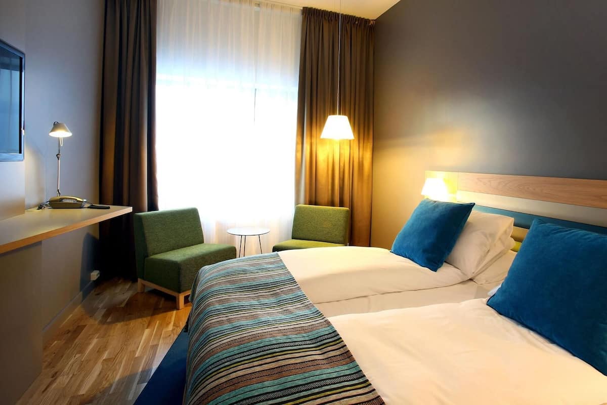 A hotel room with a bed, two chairs, a desk, a lamp, and a window with curtains.