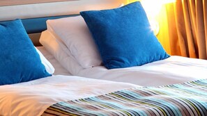 Una habitación de hotel con una cama, ropa de cama blanca y azul, una manta a rayas y un cabecero con detalles de madera.