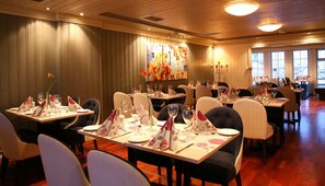 Un comedor con varias mesas dispuestas para el servicio, con piso de madera y un cuadro grande de flores en la pared.