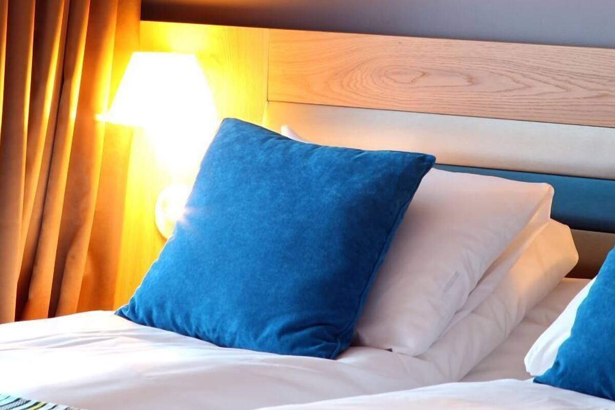 Een hotelkamer met een bed, blauwe kussens, een houten hoofdeinde en een nachtkastje met een lamp.