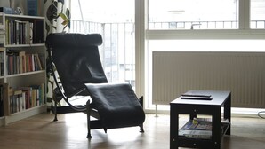Una habitación con un sillón de cuero negro, una mesita pequeña con un libro, y una estantería llena de libros.