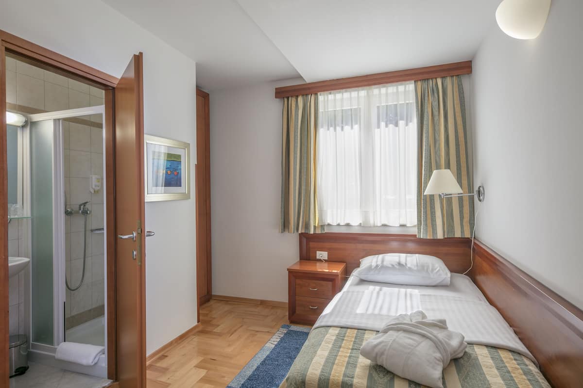 A hotel room with a wooden bed, a bedside table, a nightstand, a window with curtains, a shower area, and a framed picture on the wall.