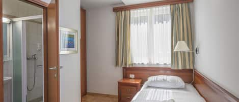 A hotel room with a wooden bed, a bedside table, a nightstand, a window with curtains, a shower area, and a framed picture on the wall.