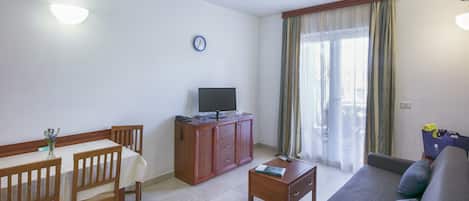 Una sala de estar con un sofá, una mesa de comedor con sillas, un televisor sobre un mueble de madera, un reloj en la pared y una ventana con cortinas.