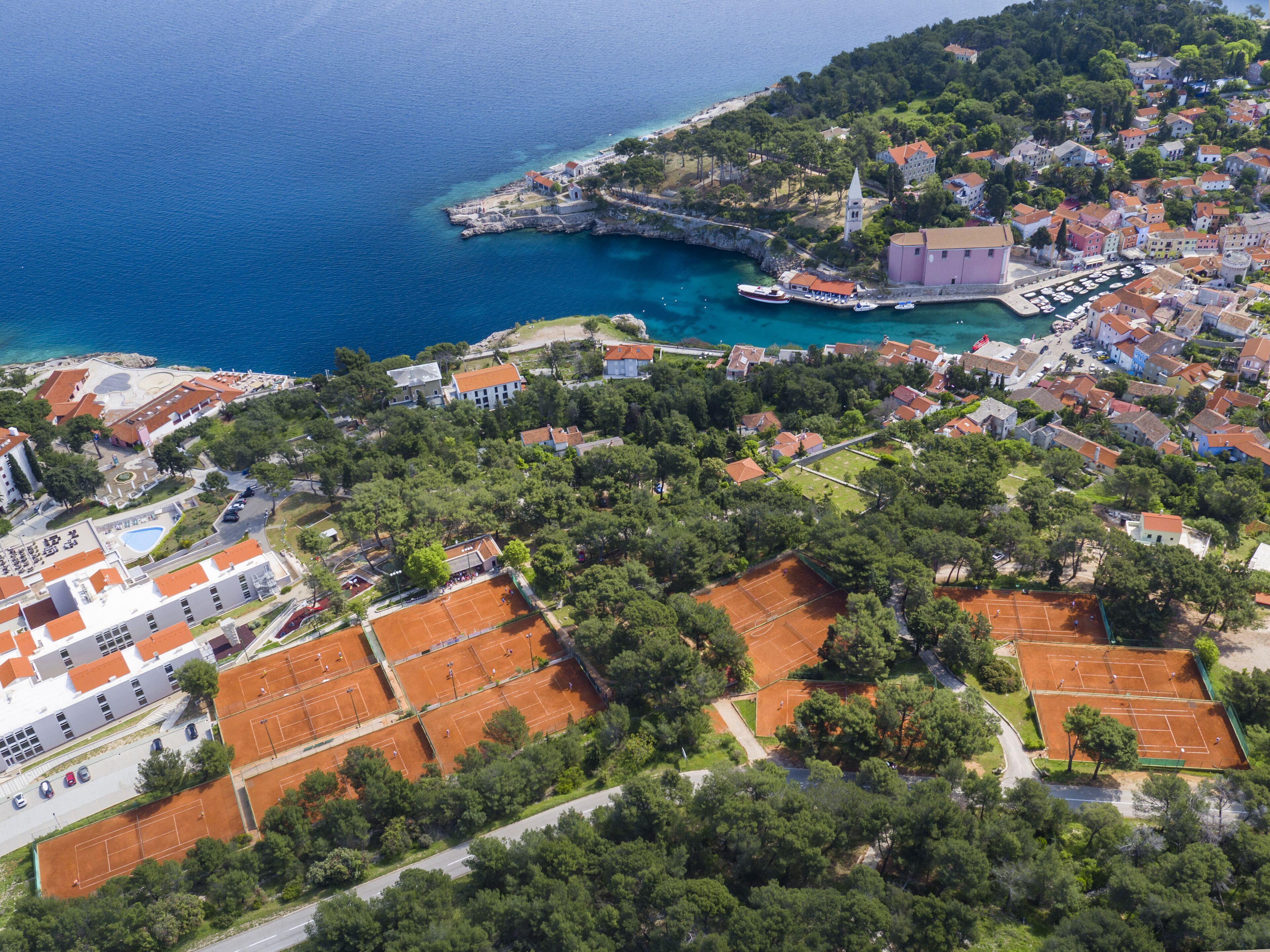 Vista aerea di una località costiera con un porto turistico, campi da tennis e edifici residenziali.